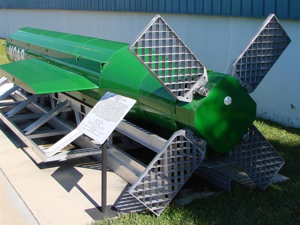 The tail fins of a large green bomb on a metal stand on grass outside a museum