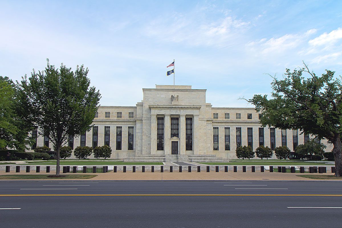 The white marble Eccles Building, headquarters of the Federal Reserve, with a U.S. flag on its roof, seen across an empty street between leafy trees