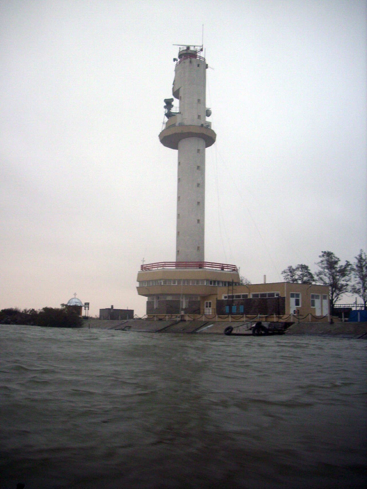 A tall white tower with a round lookout section stands on a riverbank under a gray sky, with low buildings and moored boats at its base