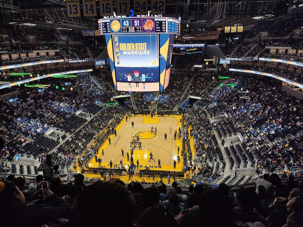 A packed indoor arena seen from the upper deck, with a large center-hung video board reading Golden State Warriors above a lit basketball court
