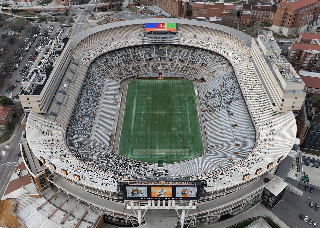 An aerial view of an empty Neyland Stadium in Knoxville, with orange-and-white checkered end zones and a scoreboard reading Neyland Stadium, Home of the Vols