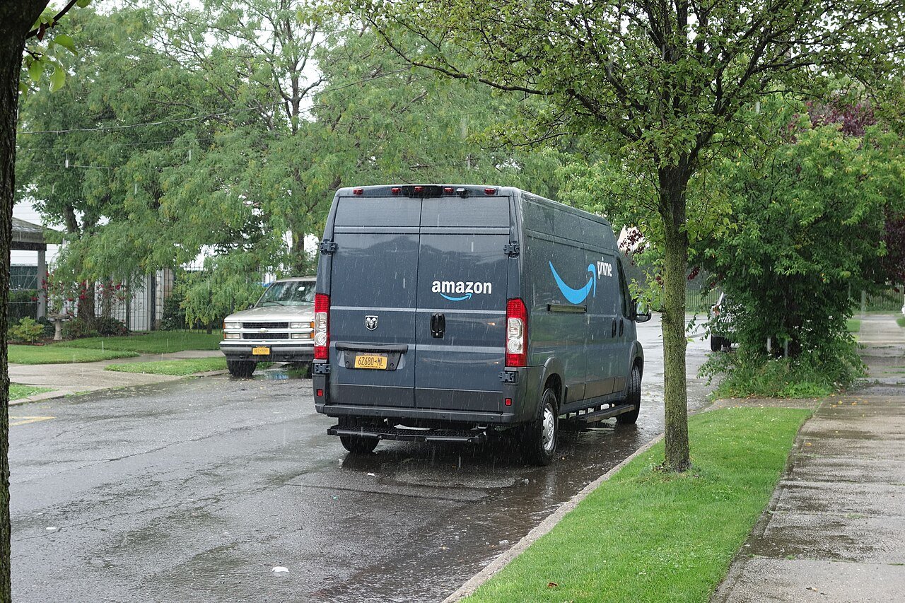 A gray Amazon Prime delivery van parked on a tree-lined residential street in Queens on a rainy day