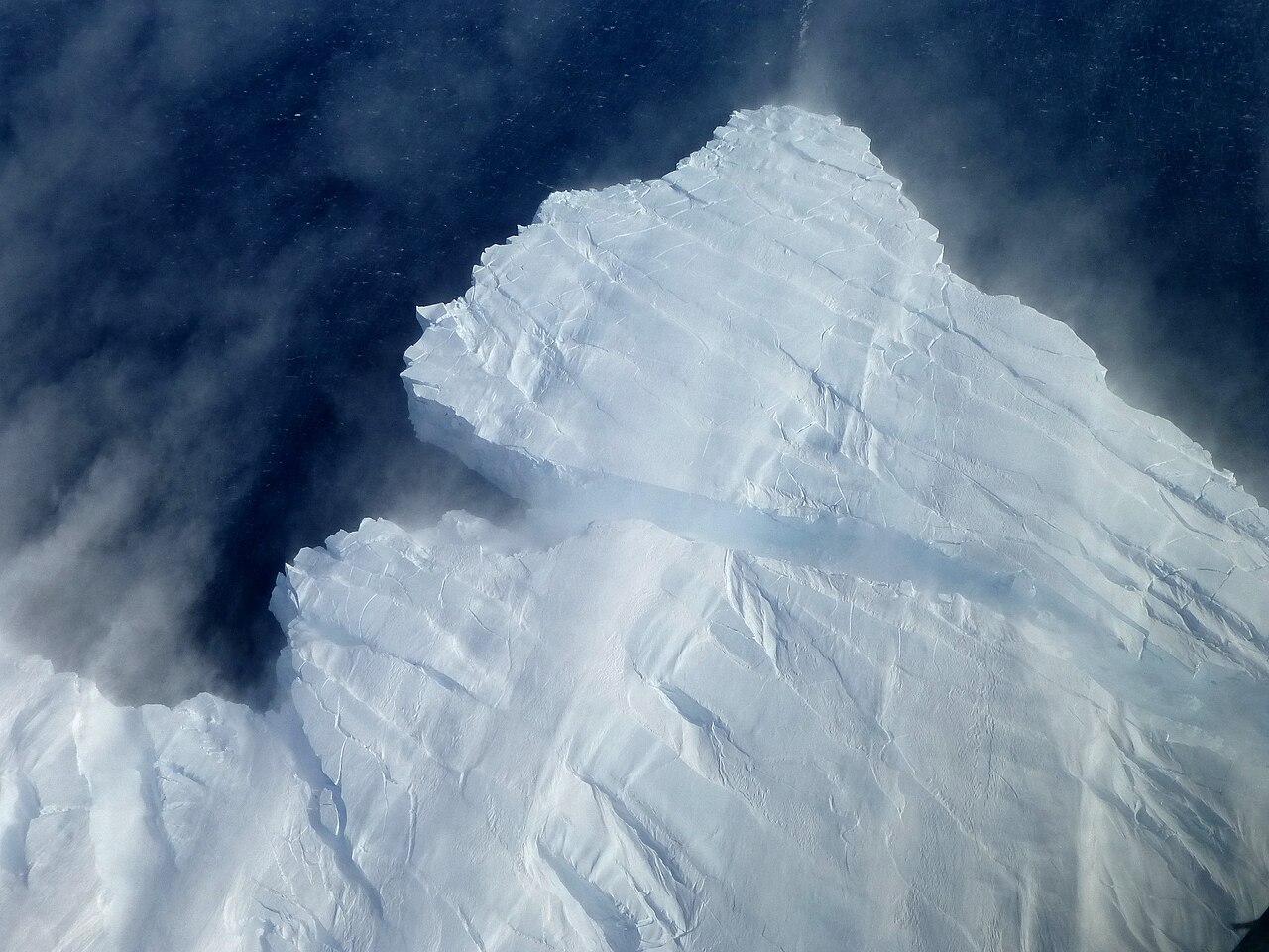 A slab of white ice breaking away from the edge of an Antarctic glacier, seen from above, with dark water and blowing snow below