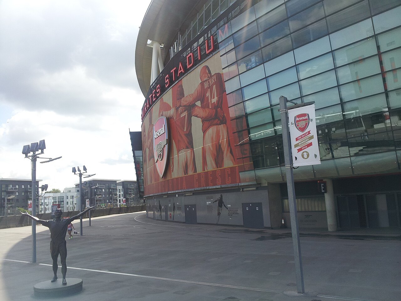 The curved glass and steel exterior of Emirates Stadium in London, with large murals of players and a statue on a plinth in the plaza
