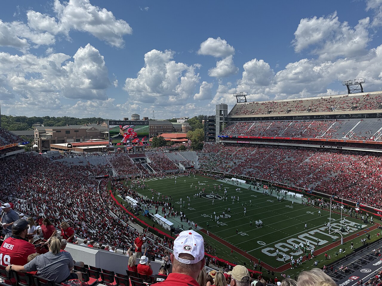 Fans in red fill the stands at Sanford Stadium on a sunny day as players in white and green line up on the field below; no Auburn players are pictured