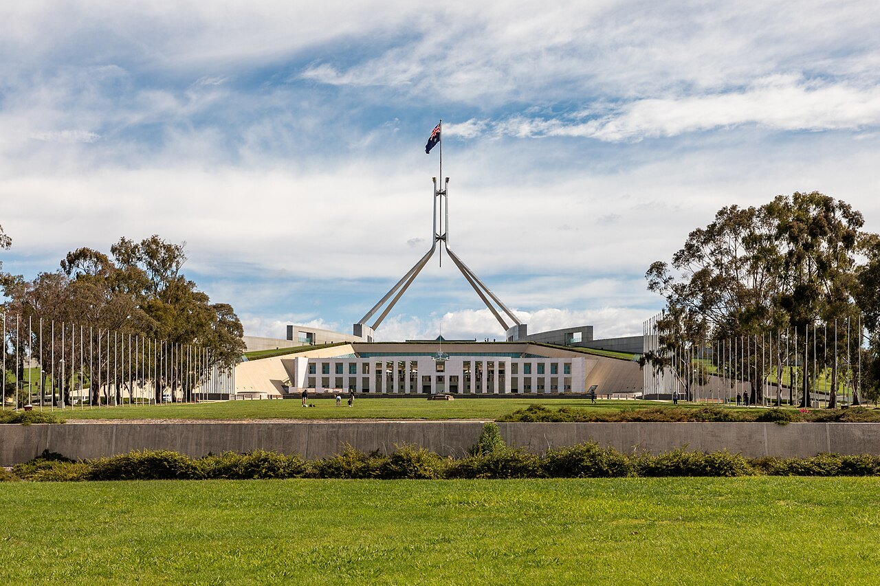 Parliament House in Canberra with its tall flagpole under a blue sky with clouds, seen across a green lawn