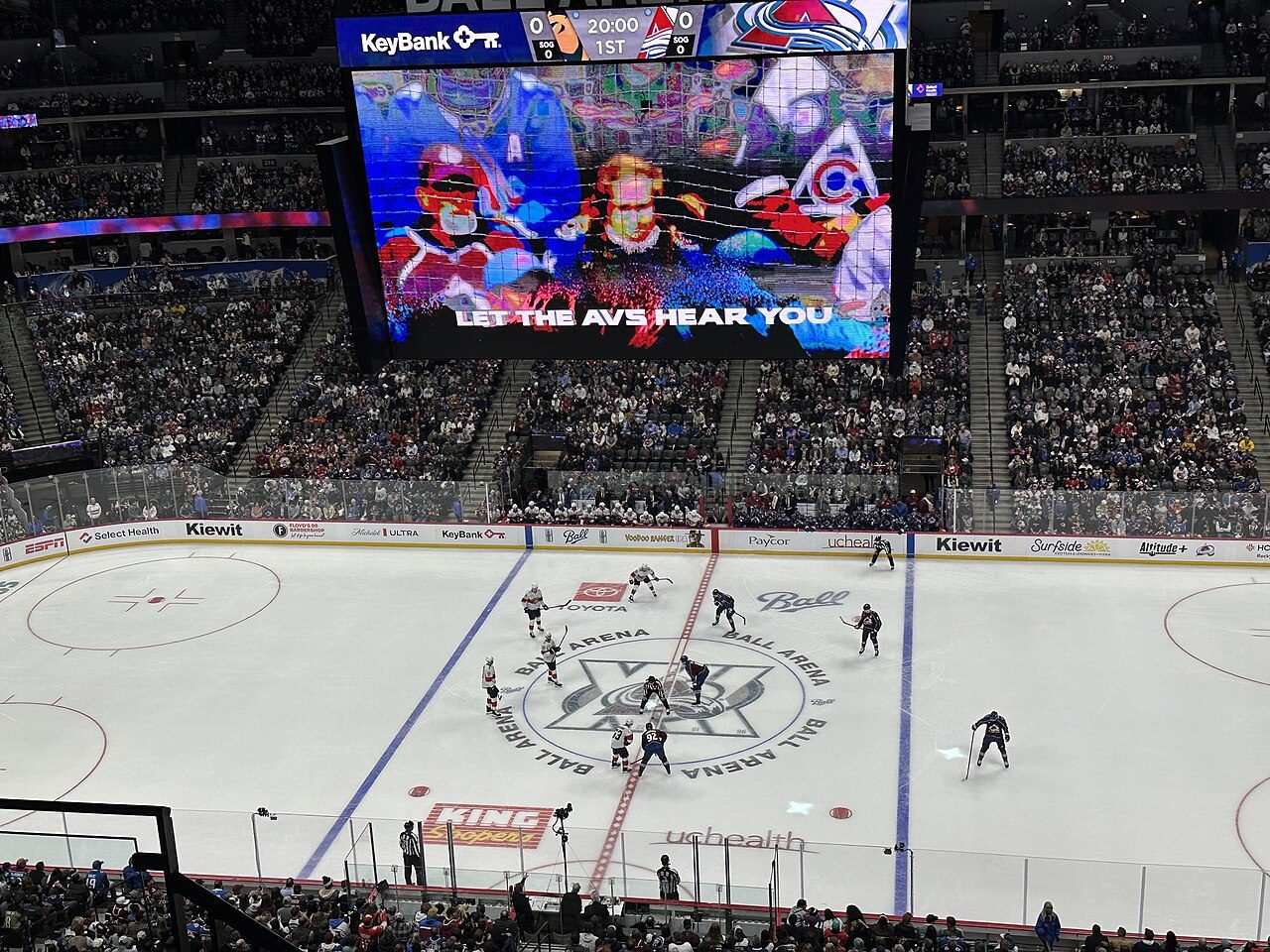 View from high in the stands of the ice at Ball Arena during a hockey game, with players at center ice and a large video board hanging above