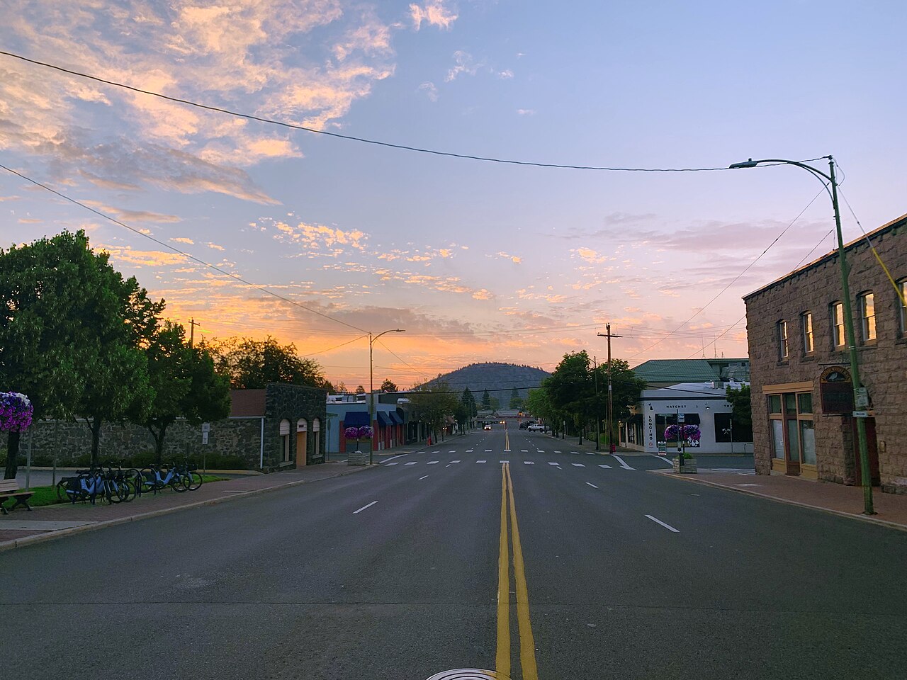 An empty downtown street in Bend, Oregon, at sunrise, with a stone building on the right and a rounded butte at the end of the road under a pink sky