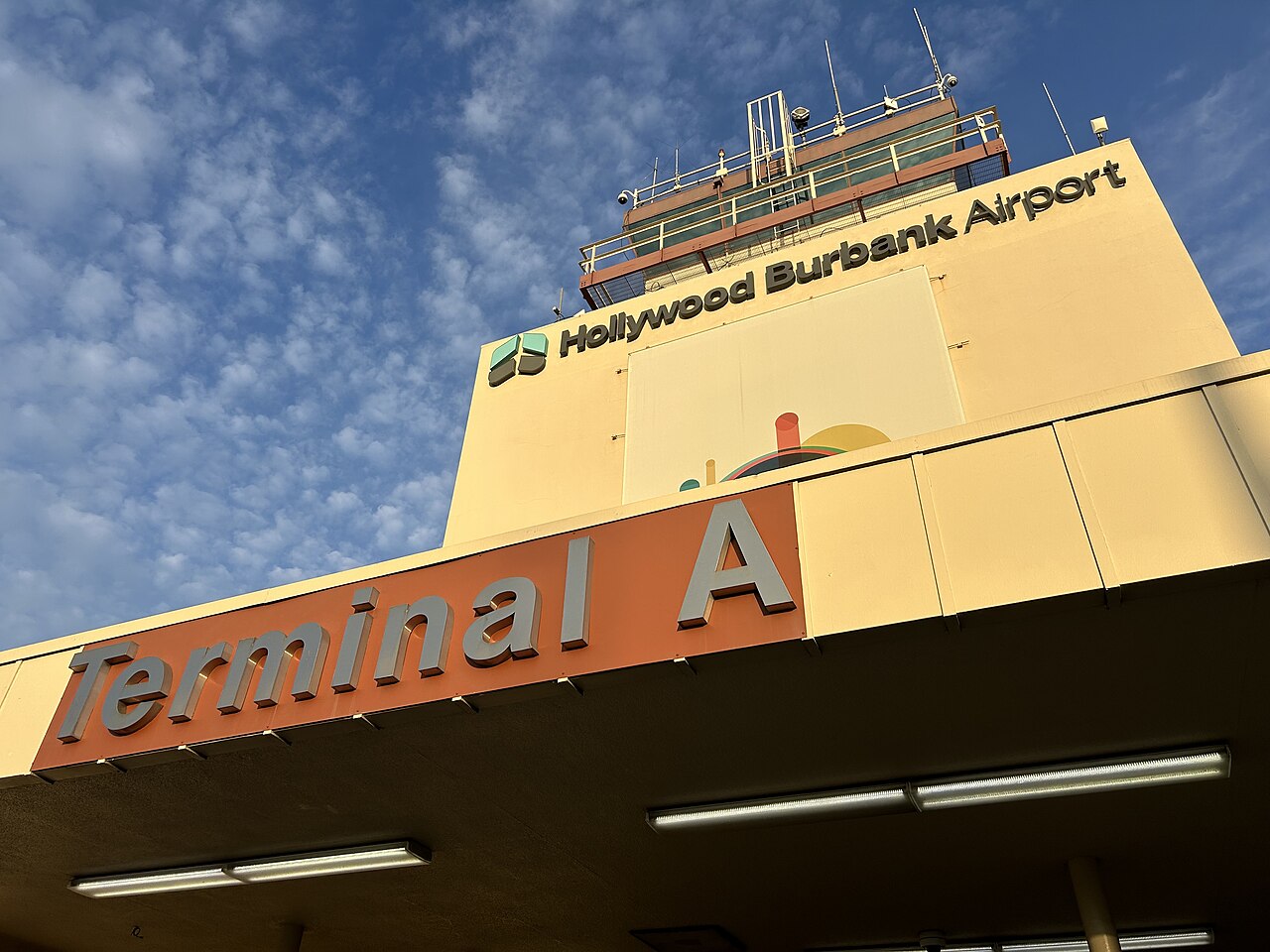 An orange Terminal A sign over the entrance of Hollywood Burbank Airport's old terminal, with the airport's name on a tan tower above