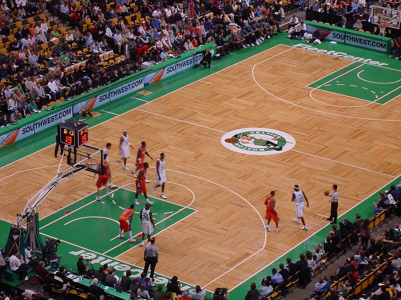 View from high in the stands of a Celtics home game, with players in white and orange jerseys on a wooden parquet court, the Celtics logo at center court and fans in the seats