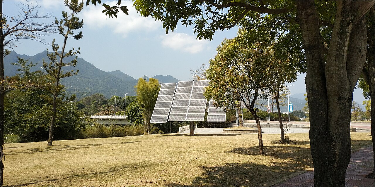 Tilted solar panels mounted on poles stand on a dry grassy lawn among trees, with a road bridge, streetlights and green mountains behind them under a partly cloudy sky
