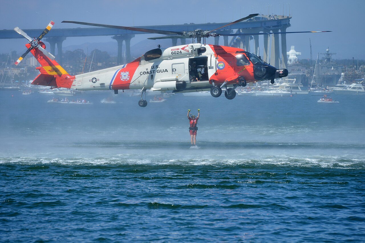 An orange and white U.S. Coast Guard MH-60T Jayhawk helicopter hovers over San Diego Bay as a rescue swimmer hangs on a cable above the water