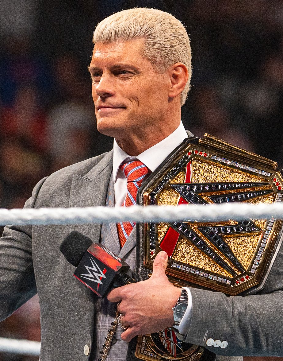 Cody Rhodes in a gray suit holds a microphone and a championship belt at ringside