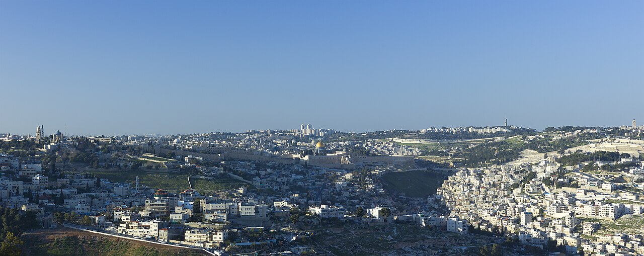 A wide view of Jerusalem's Old City and surrounding neighborhoods under a clear blue sky, seen from a hillside promenade