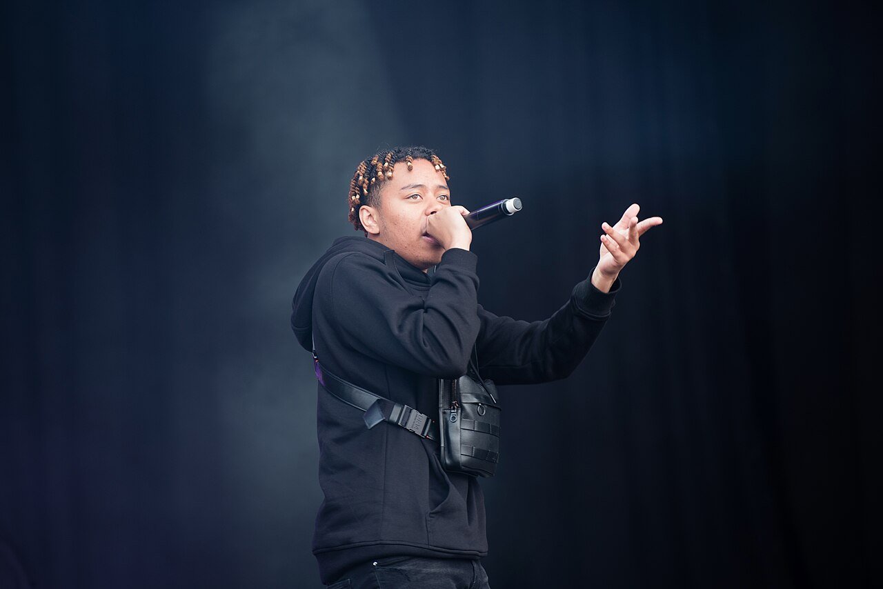 Rapper Cordae in a black hoodie performs on stage, holding a microphone and pointing toward the crowd