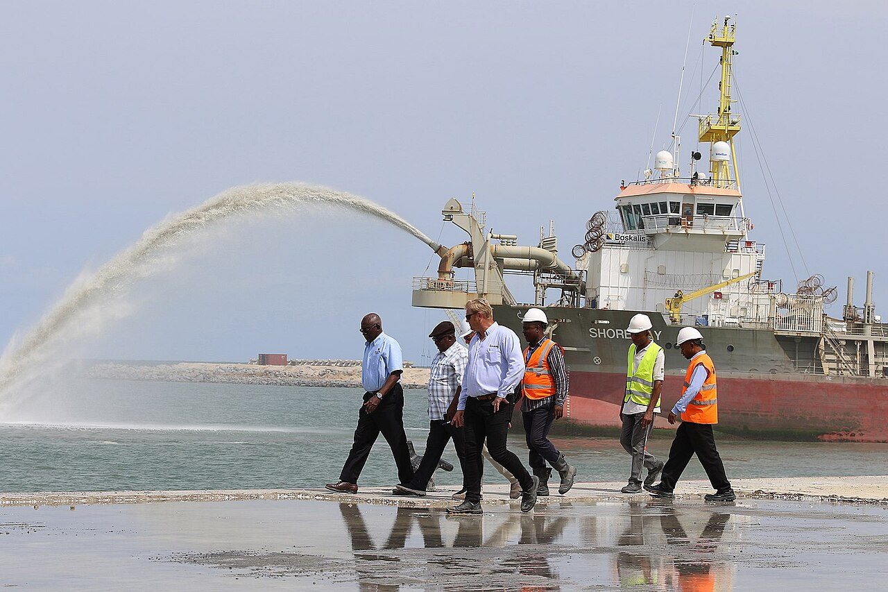 A dredging ship sprays a long arc of sand and water near the shore as seven people walk across wet sand in the foreground, six men clearly visible and a seventh person mostly hidden behind them, four in white hard hats and three in orange or yellow safety vests