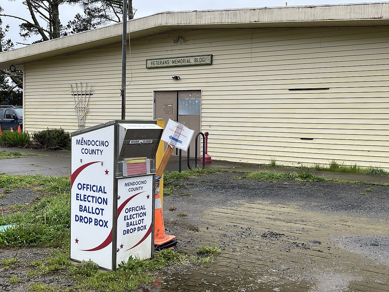 A white official election ballot drop box with a red stripe, labeled Mendocino County, standing on a post outside a pale yellow wooden building