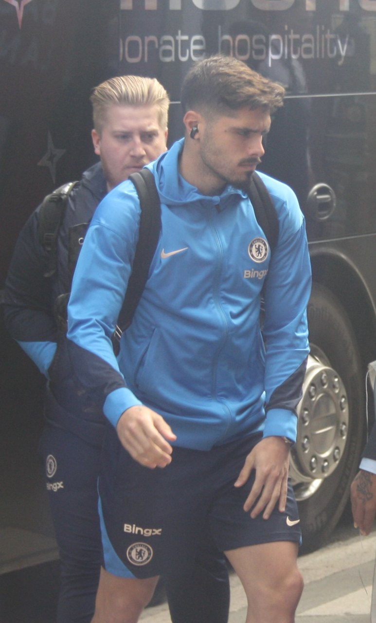 Pedro Neto in a blue Chelsea training top and shorts, carrying a backpack, with another person behind him next to a team bus