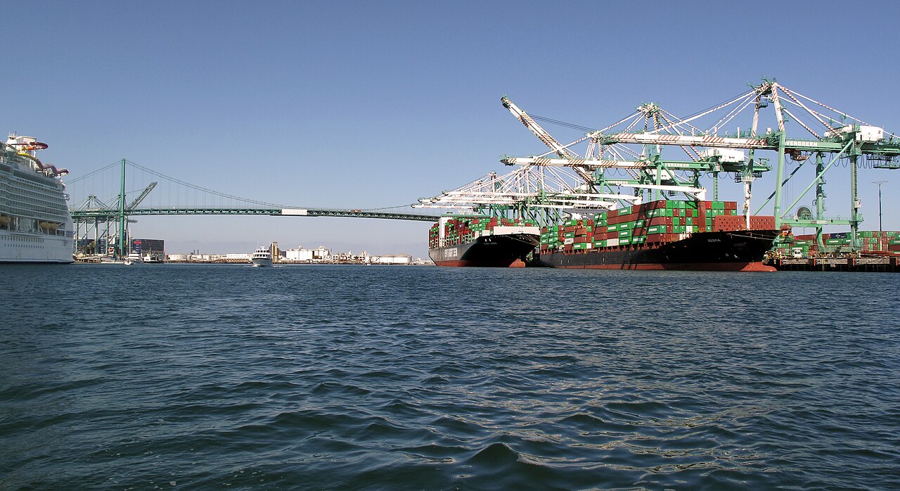 Two container ships stacked with colorful containers under large green cranes at the Port of Los Angeles, with a suspension bridge in the background