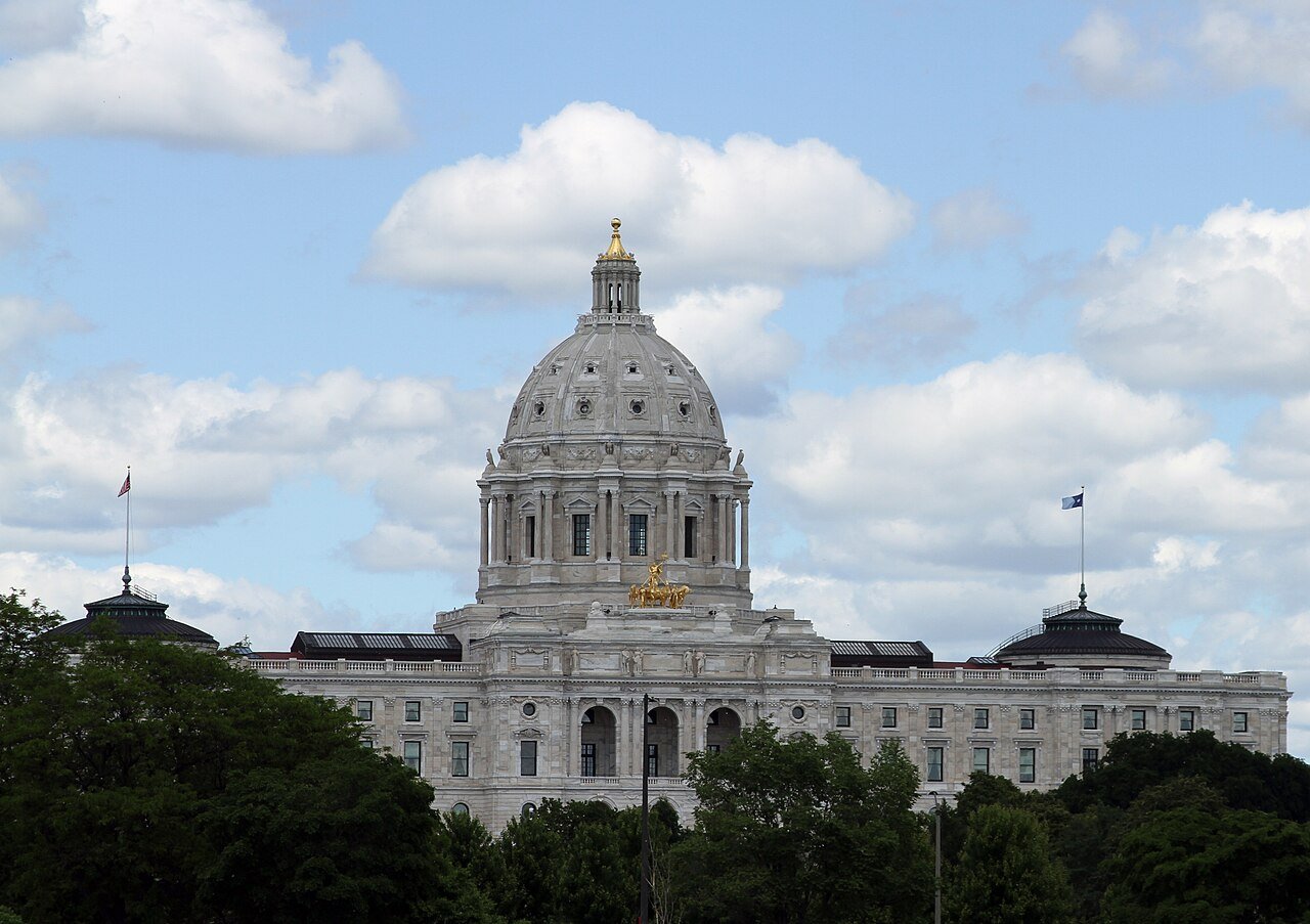 The white marble dome of the Minnesota State Capitol in St. Paul under a blue sky with clouds, with trees in the foreground
