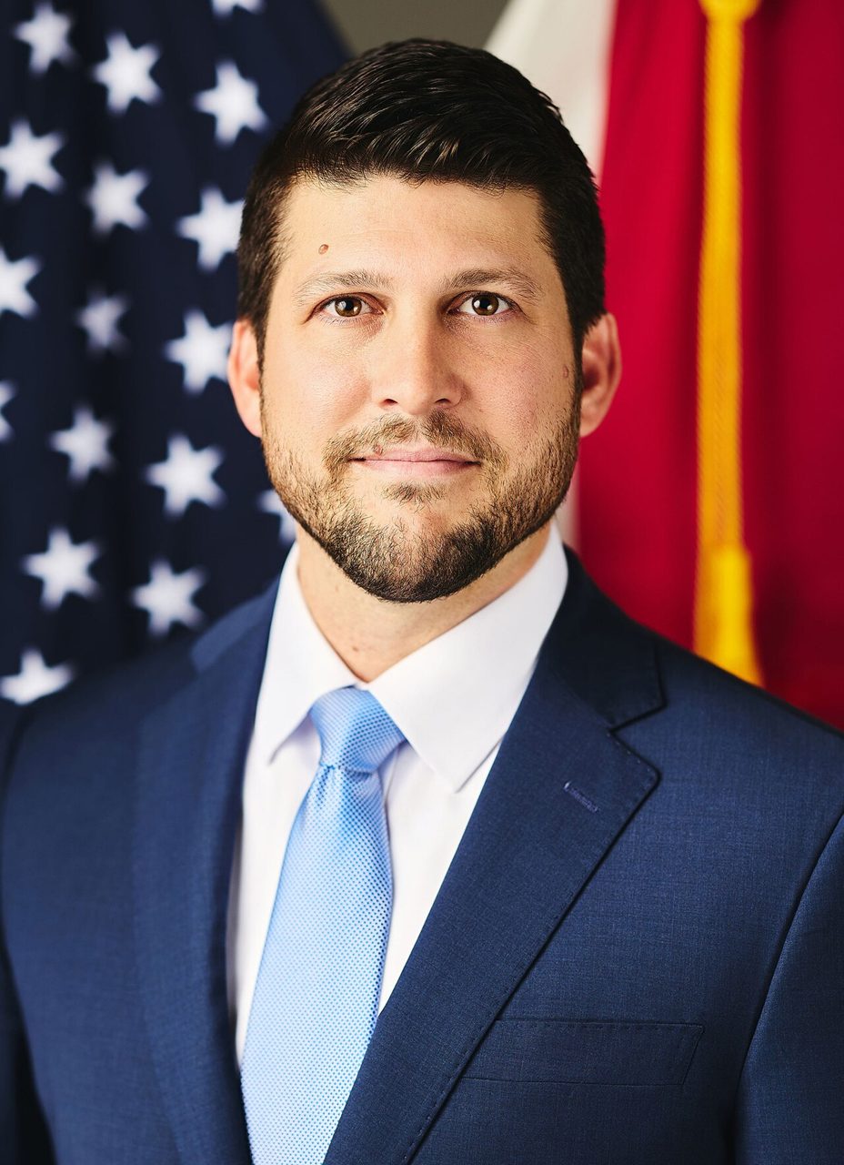 Official portrait of Florida Attorney General James Uthmeier, who filed the suit against Discord, in a dark suit and light blue tie in front of U.S. and Florida flags; Discord disputes the suit