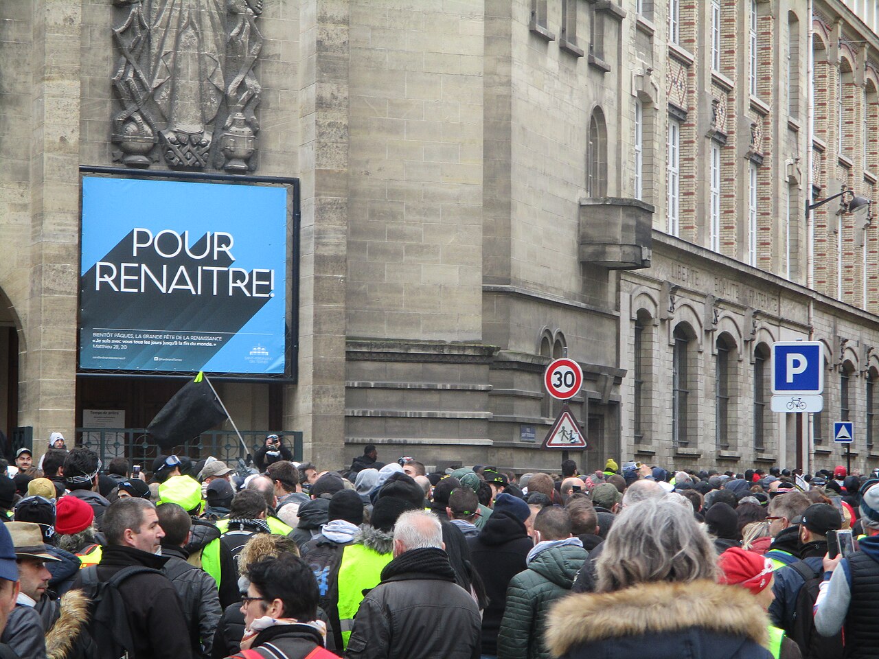 A crowd of protesters, some in yellow vests and one holding a black flag, fills a Paris street beside a stone church in 2019