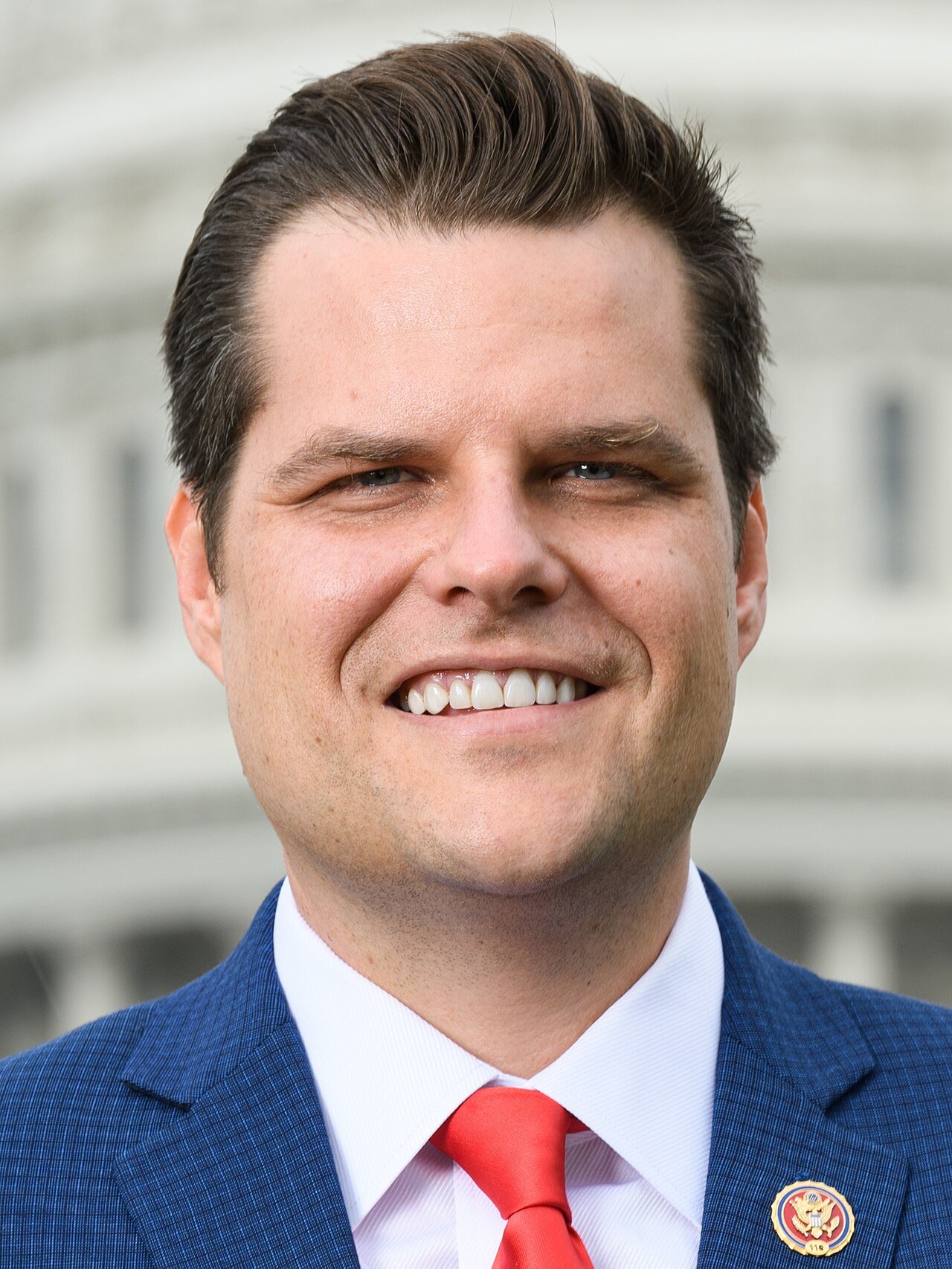 Official portrait of Matt Gaetz in a dark suit and red tie, smiling in front of the U.S. Capitol dome
