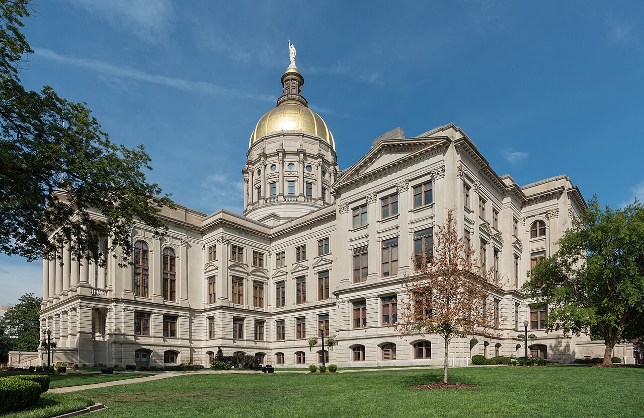 The Georgia State Capitol in Atlanta, a white stone building with a gold dome and columned wings, seen from the west across a green lawn