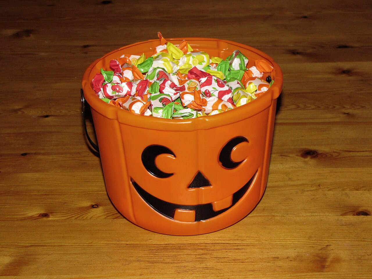 An orange plastic jack-o'-lantern bucket filled with colorful wrapped hard candies, sitting on a wooden floor