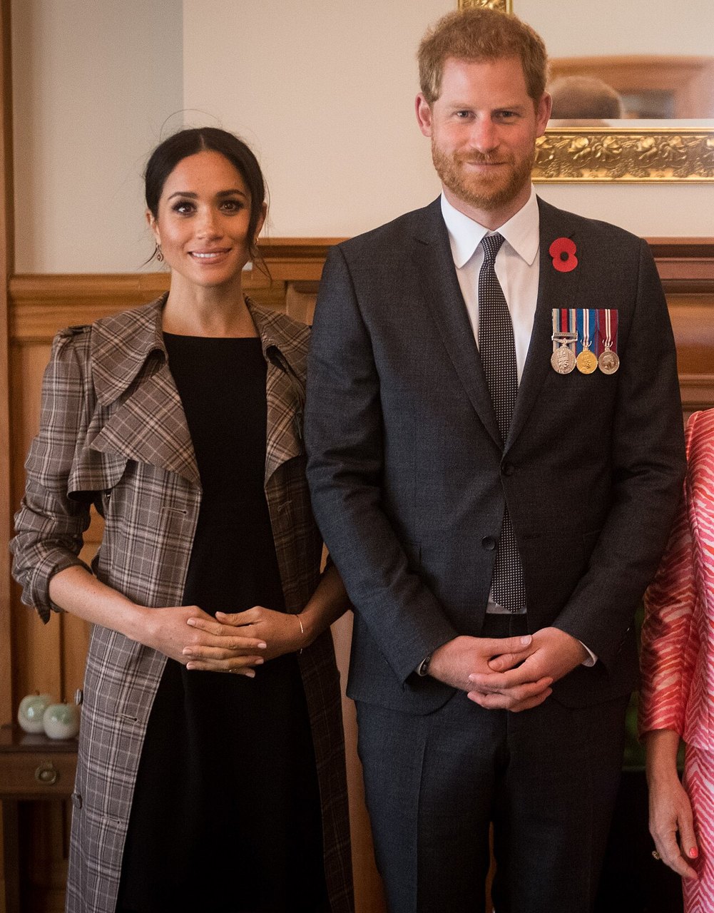 Meghan in a plaid coat and Prince Harry in a dark suit with medals and a poppy, standing side by side indoors, with part of another person at the right edge