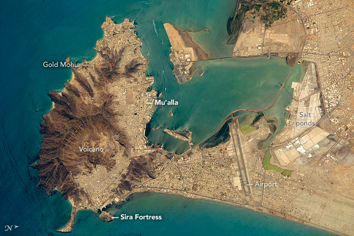 An annotated astronaut photograph of Aden, Yemen, from space, showing a brown volcanic peninsula, a turquoise bay, the port, the airport and salt ponds