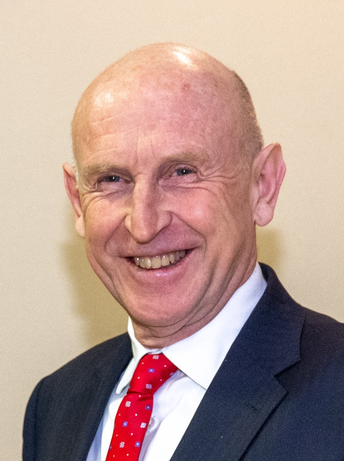 John Healey, smiling, in a dark suit, white shirt and red patterned tie against a cream wall