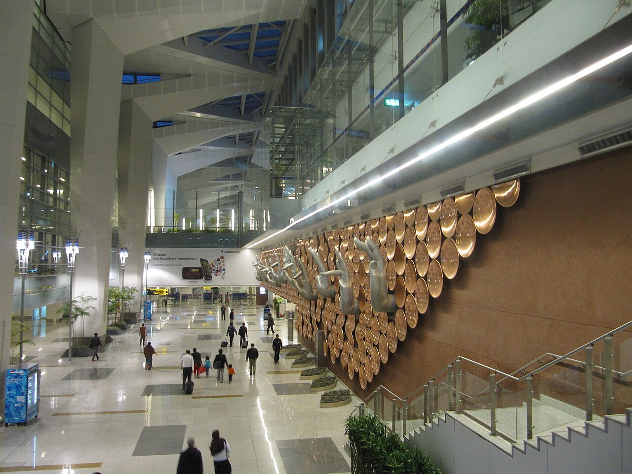 Travelers walk through the bright main hall of an airport terminal, past a wall decorated with large copper-colored discs