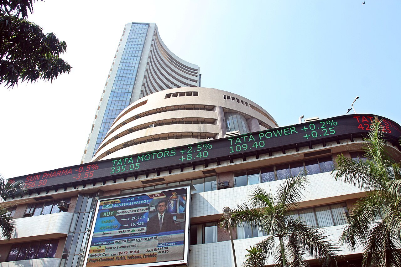 A tall modern tower rising behind a curved building with an electronic ticker board, framed by palm trees under a pale sky