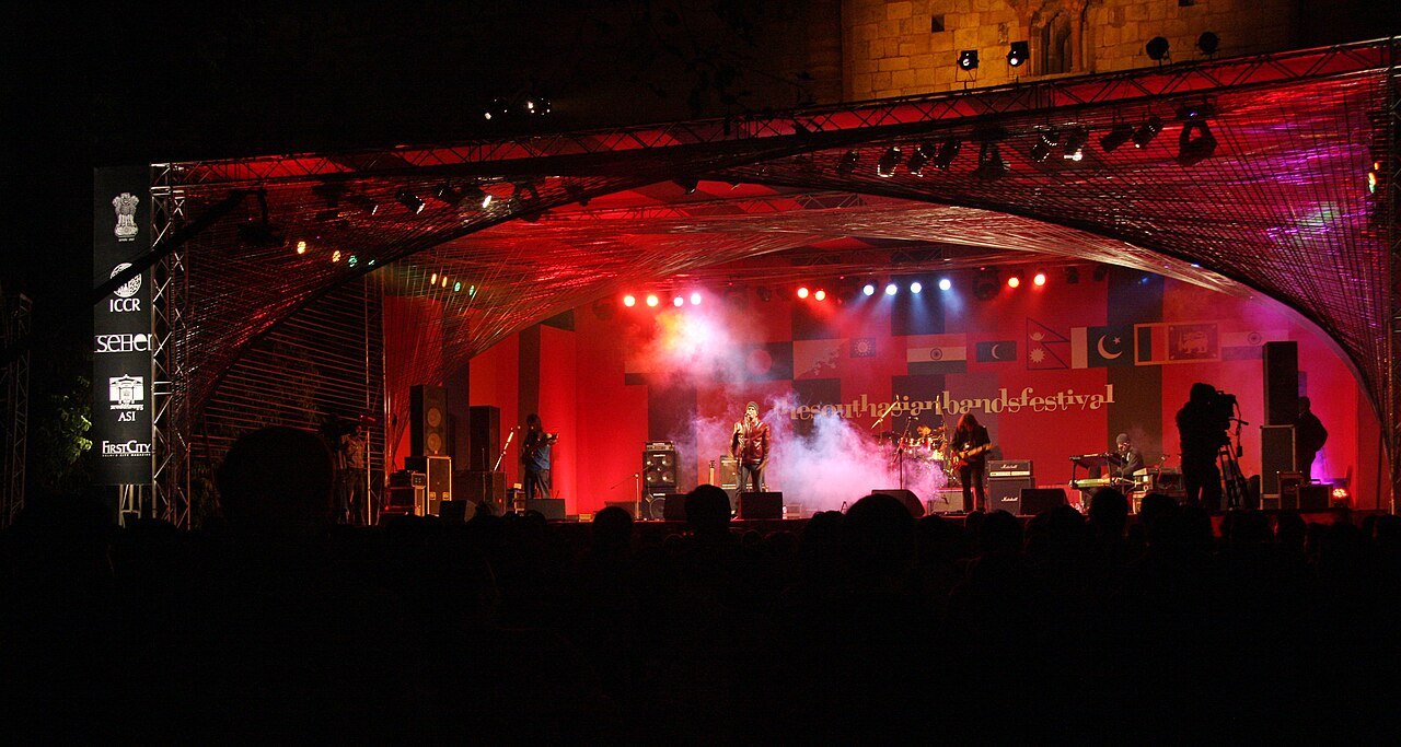 A rock band performs on a stage lit in red under a large arched canopy at night