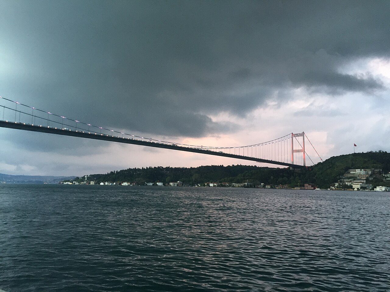 Dark storm clouds over the Bosphorus and the Fatih Sultan Mehmet Bridge in Istanbul, with wooded hills and houses along the shore