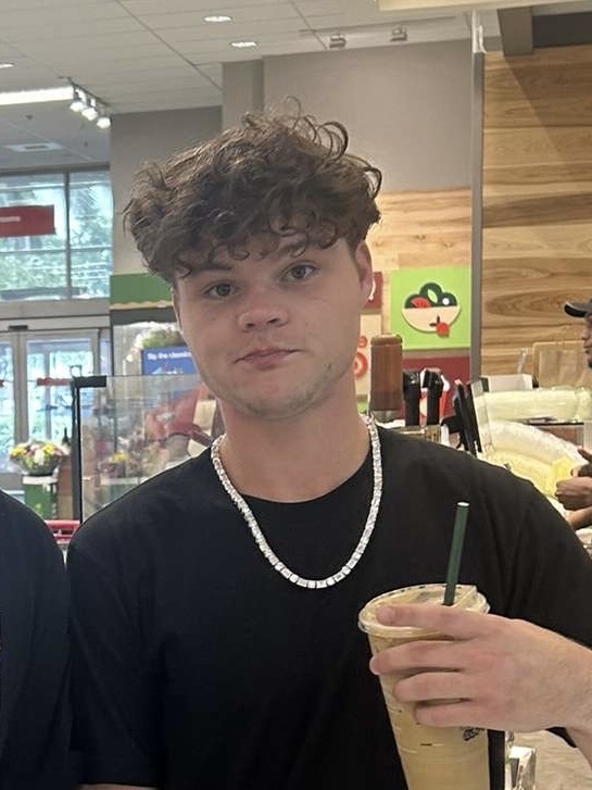 Jack Doherty, with curly brown hair and a silver chain, holds an iced drink inside a store