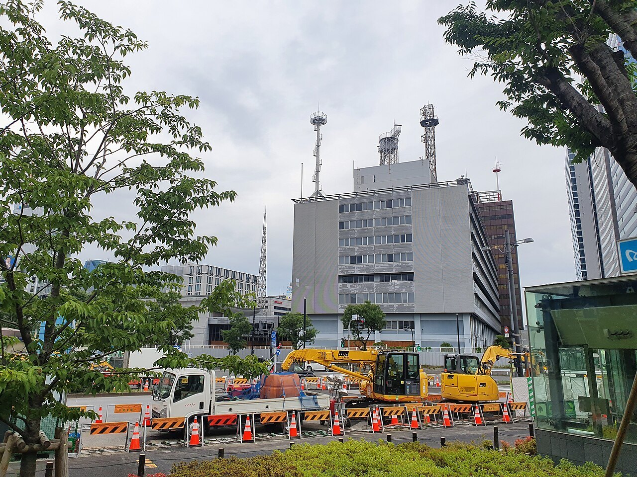 A multistory office building with antennas on the roof behind a busy street, with construction barriers and trees in the foreground