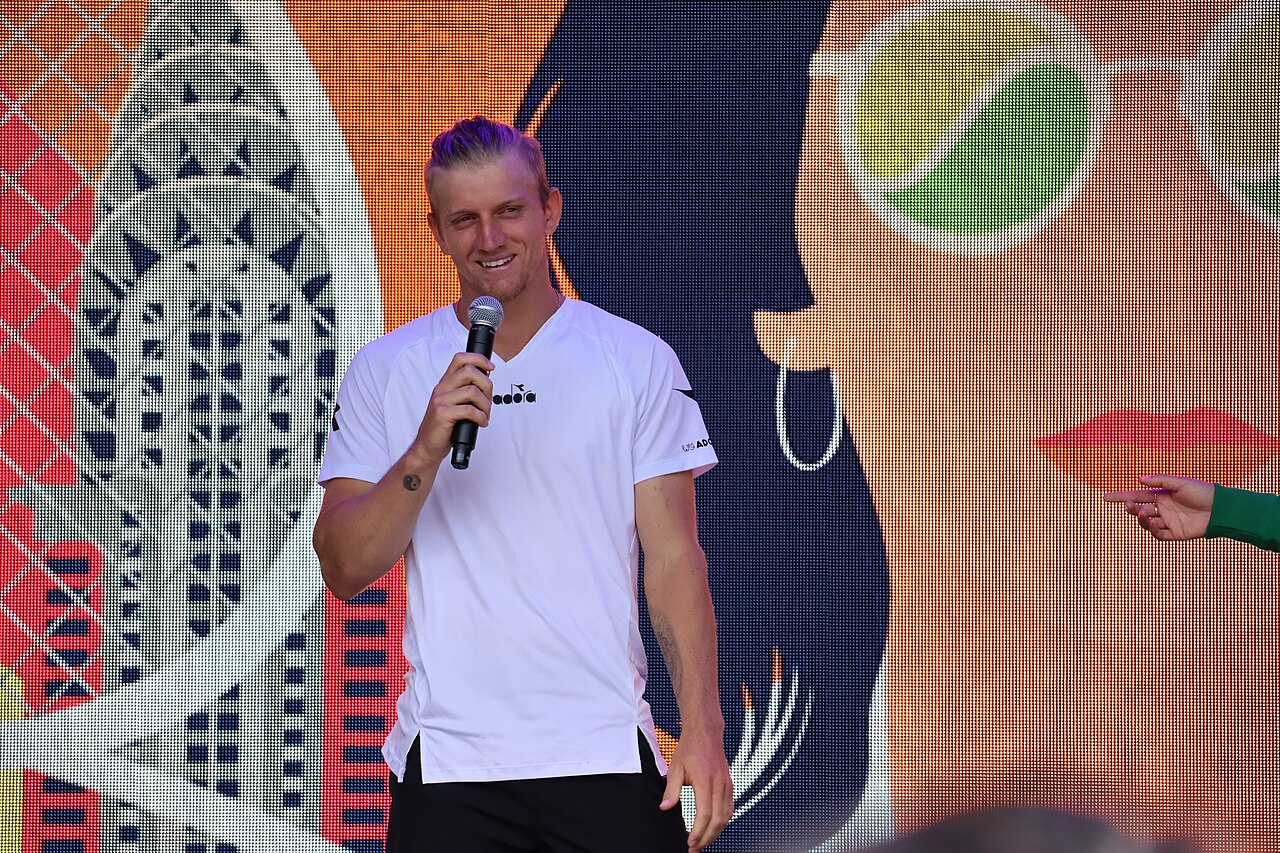 Alejandro Davidovich Fokina, in a white shirt, smiles while holding a microphone on a stage in front of a colorful mural