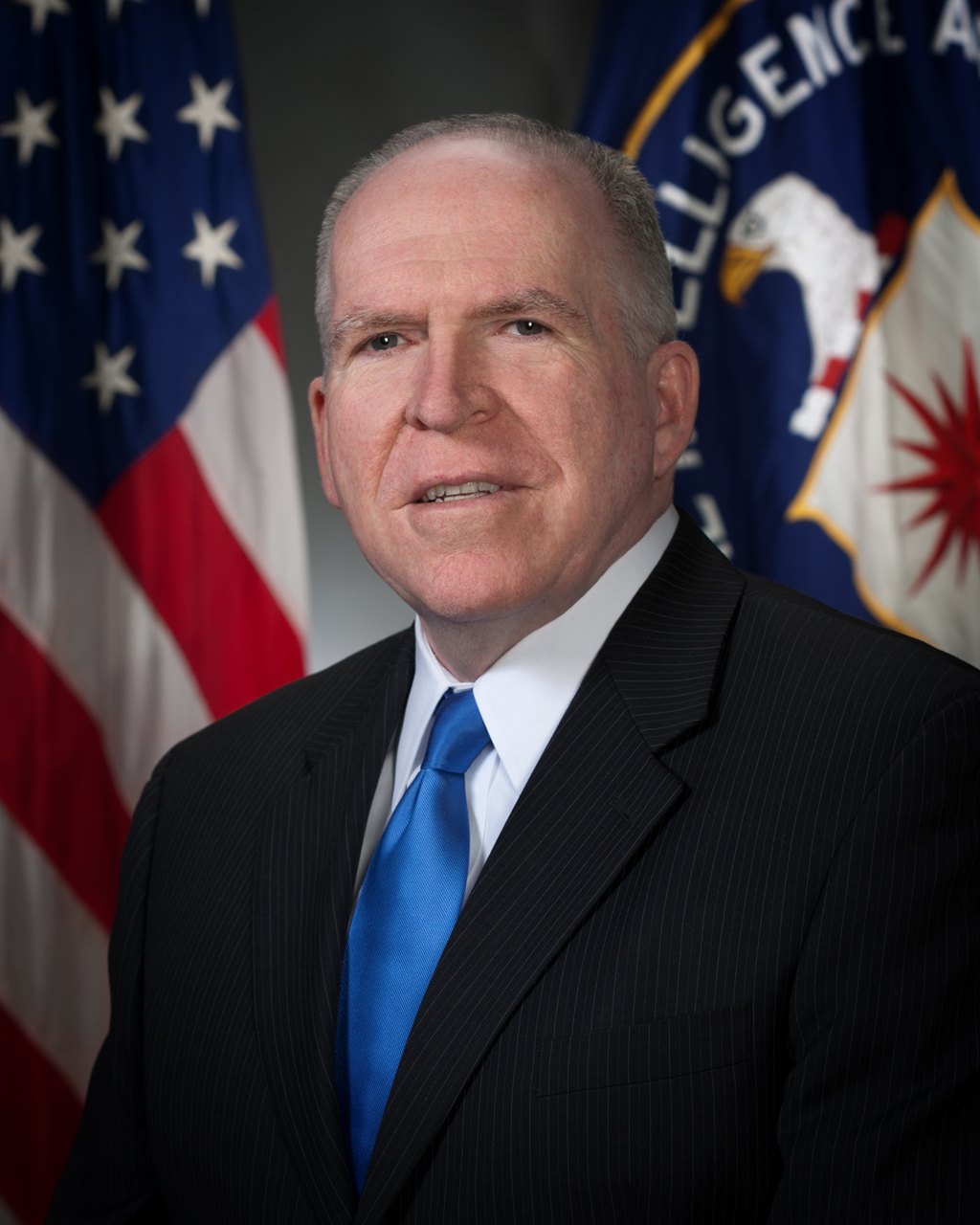 Official portrait of John Brennan in a dark suit and blue tie, standing before a U.S. flag and the CIA flag