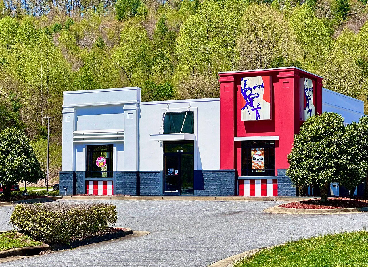 A KFC restaurant with a white and red facade, a Colonel Sanders logo and red-and-white striped trim, surrounded by trees and a parking lot