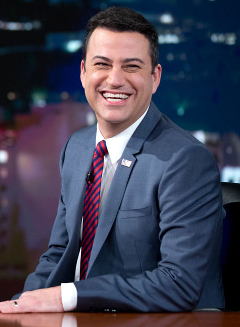 Jimmy Kimmel, smiling, in a blue suit, white shirt and red striped tie, seated at a desk on a television set