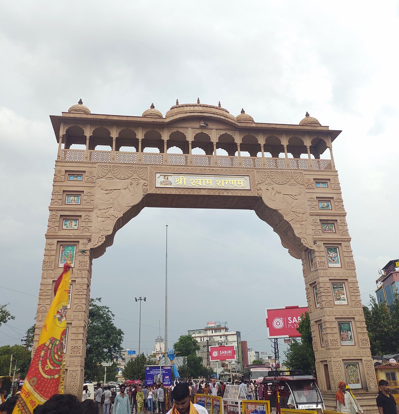 A tall carved sandstone arch over a busy street with crowds of people and saffron flags, marking the entrance to the temple town of Khatu Shyam Ji