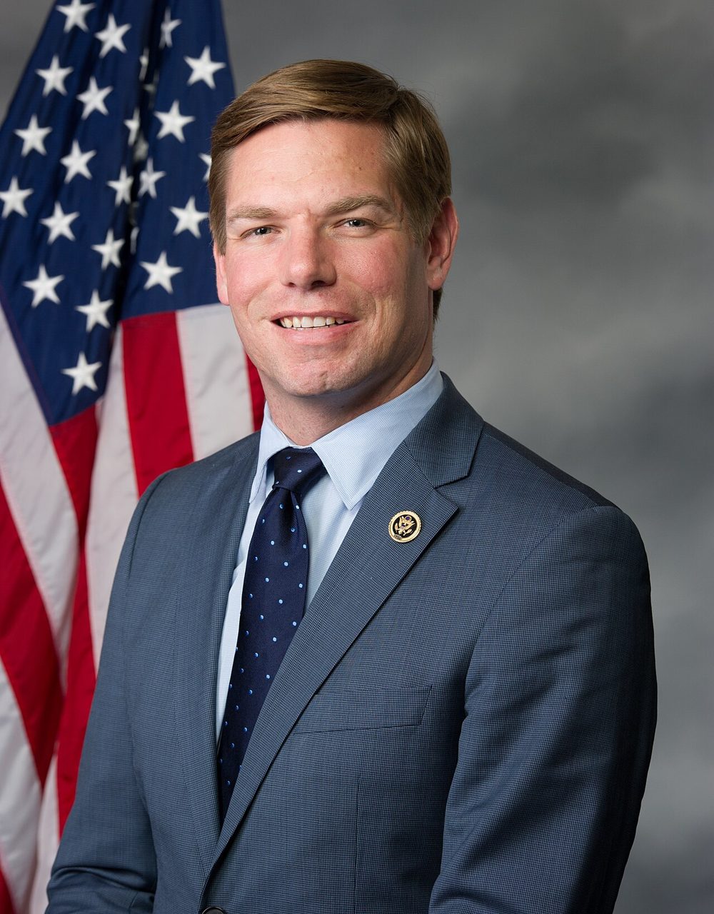 Official portrait of Eric Swalwell smiling in a blue suit, light blue shirt and navy polka-dot tie, in front of an American flag and a gray backdrop