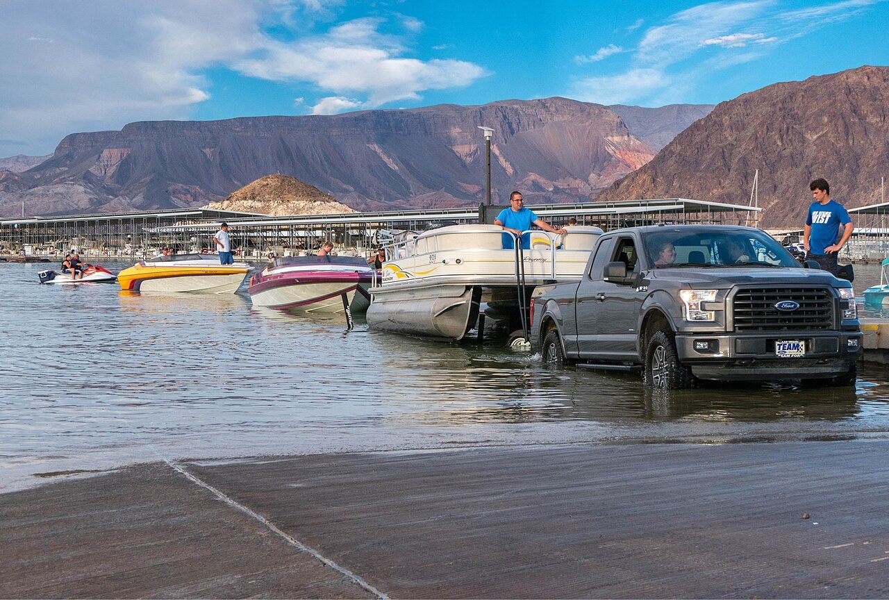 A pickup truck and several boats at a concrete launch ramp on Lake Mead, with people on the boats and rugged mountains under a blue sky behind