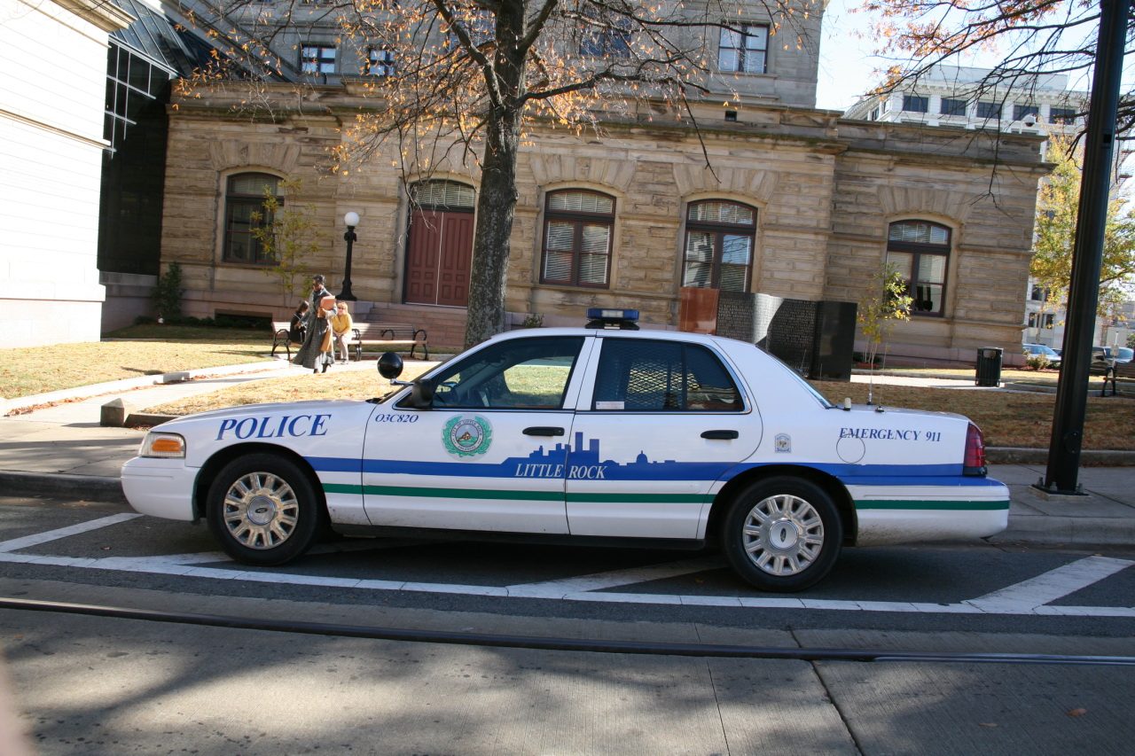 A white Little Rock police patrol car with a blue stripe parked on a street in front of an older brick building with tall windows