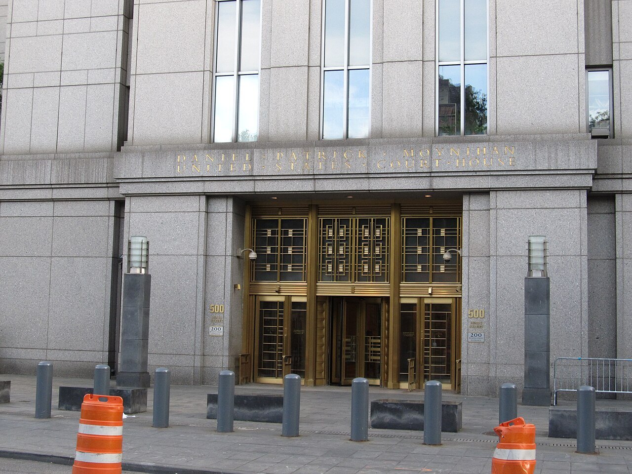 The granite entrance of the Daniel Patrick Moynihan U.S. Courthouse in Manhattan, with bronze doors, bollards and two orange traffic barrels