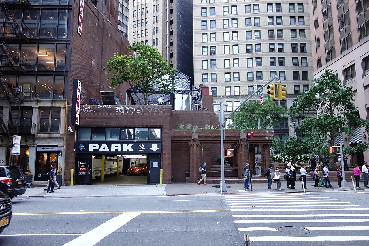 The entrance to a 24-hour parking garage under an office building in Lower Manhattan, with a large PARK sign and pedestrians on the sidewalk