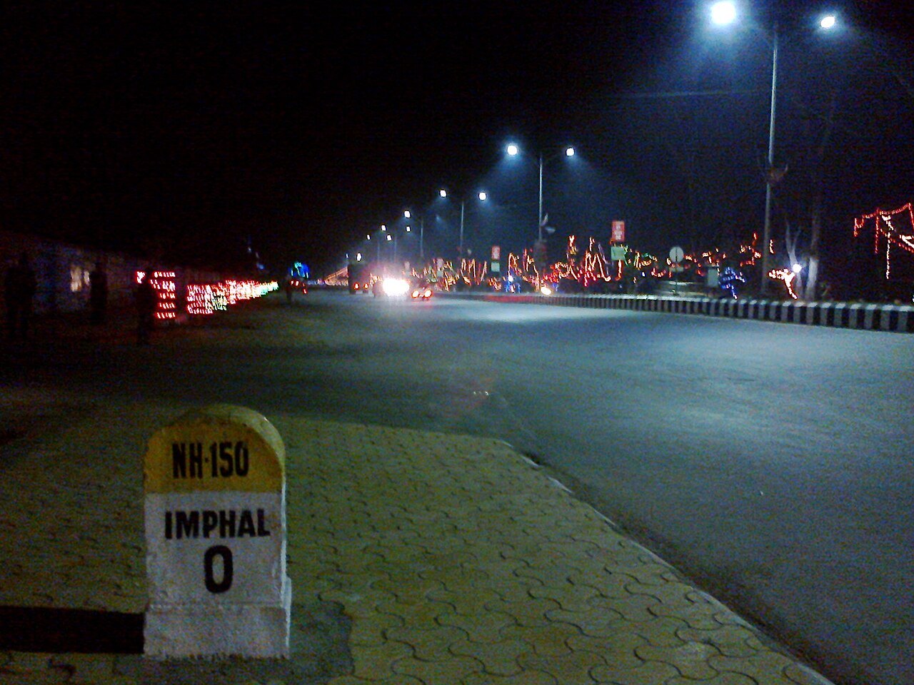 A night street scene with a roadside milestone reading "NH-150 Imphal 0" in the foreground and street lights and colored lights along the road
