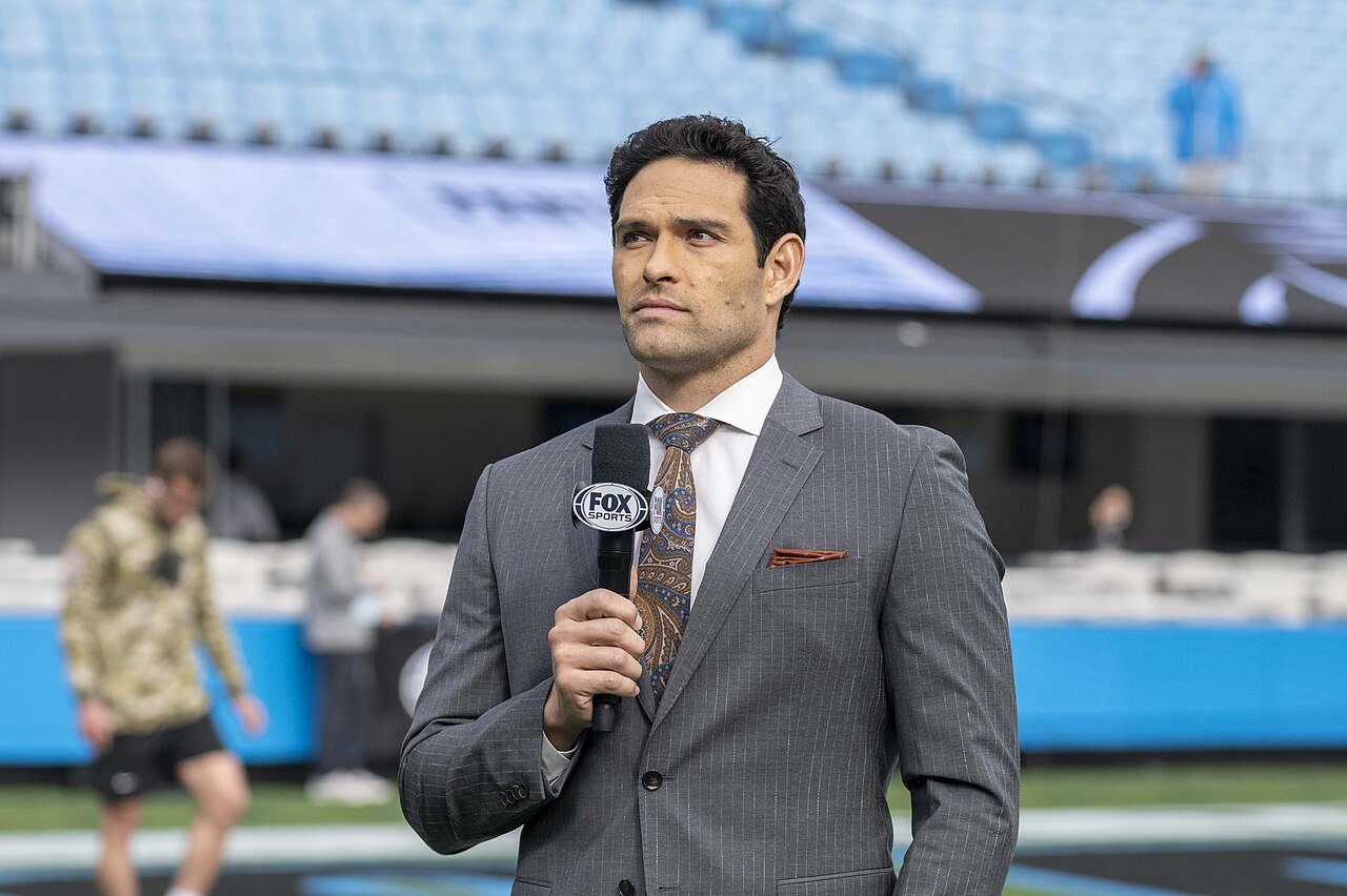 Mark Sanchez in a gray suit holds a FOX Sports microphone on the field of an empty football stadium